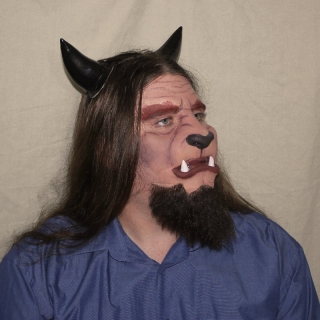 A man wearing a foam latex beast face prosthetic with makeup, beard and horns.