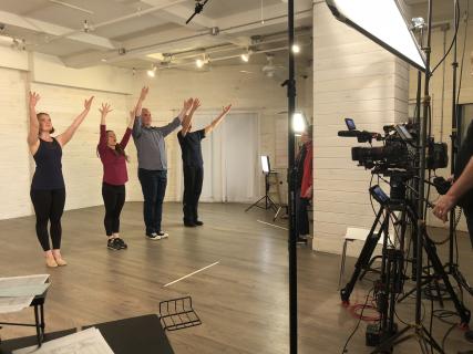 Four dancers stand in a row with their arms in their air and feet together in front of a filming camera.