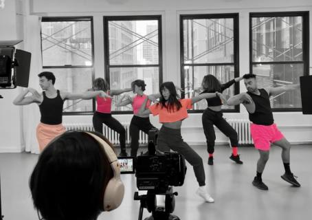 Five dancers wearing pink tops and shorts in a row demonstrating a dance move while someone is filming in front of them.