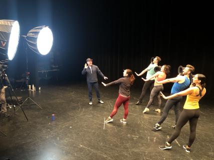 A choreographer demonstrating a move as four dancers behind perform the movement in front of bright lights.