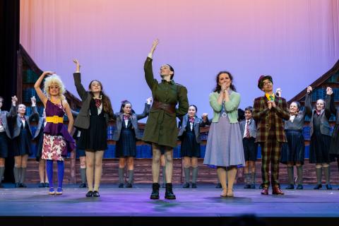The lead cast of Matilda the Musical posed on stage in costume, Miss Trunchbull in the middle, flanked by Miss Honey, Matilda, Ms. Wormwood and Mr. Wormwood, with supporting characters smiling in the background.