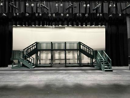 Empty stage with multi-level wooden Newsies set featuring central platform and stairs on both sides.